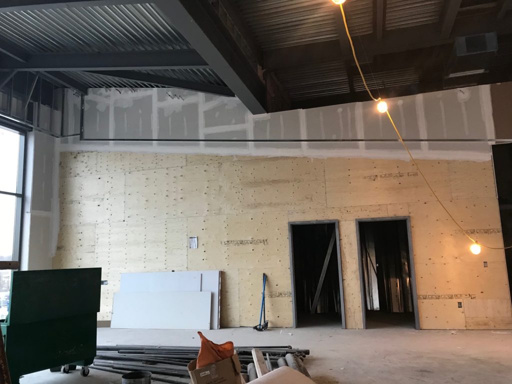 Interior picture of construction of the community room in the police headquarters building. It shows plywood and drywall on the walls as well as some resting against the wall, ready to be used. The image is filled with natural light, coming in through large windows. Other construction and building materials appear, such as steel beams and plumbing material ready to be installed.