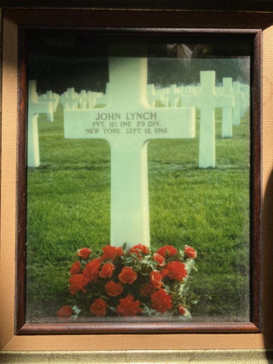 A colour photograph of a colour photograph of a cross at a gravesite with red flowers at its base. The cross reads, “JOHN LYNCH PVT 115 INF. 29 DIV NEW YORK SEPT 18, 1918.”