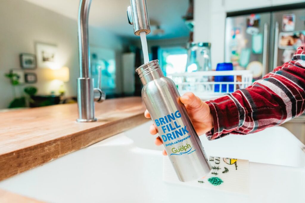 A hand holding a Guelph-branded metal water bottle and filling it with tap water from a kitchen sink. The bottle has text reading "Bring, fill, drink, guelph.ca/tapwater."