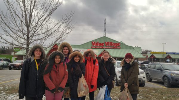 A colour photograph of seven people huddled together in front of Value Village. They are wearing jackets, many with their hoods up, and carrying plastic bags.