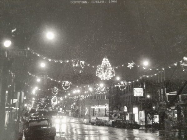 An old, black and white photograph of a street at night, with decorative lights strung across it. At the top it reads, DOWNTOWN, GUELPH, 1966. 