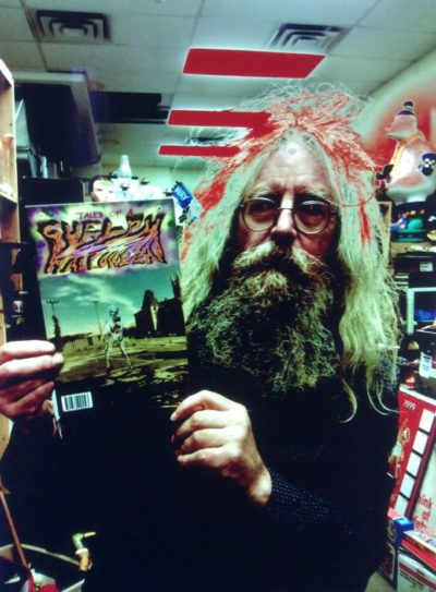 A colour photograph featuring an older man with long hair and a long beard posing with a comic book. He wears glasses, is dressed in black, and doesn’t smile.
