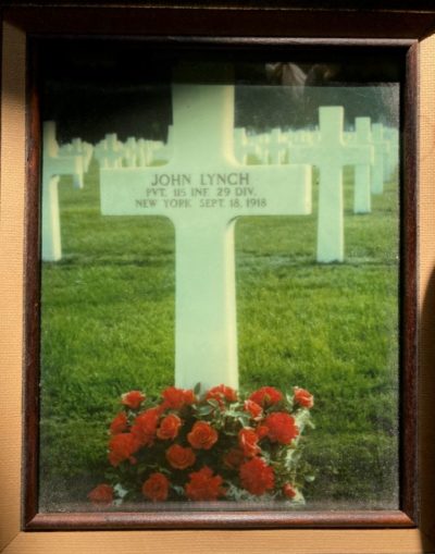 A colour photograph of a colour photograph of a cross at a gravesite with red flowers at its base. The cross reads, “JOHN LYNCH PVT 115 INF. 29 DIV NEW YORK SEPT 18, 1918."