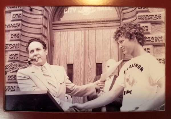 An old, sepia tinted photograph with former Guelph Mayor, Norm Jary, at a podium in front of City Hall, shaking the hand of Terry Fox, who wears a “Marathon of Hope,” t-shirt.