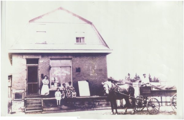 A black and white photograph. Four people are on a porch, one stands and three sit. A man sits in his wagon attached to a horse.