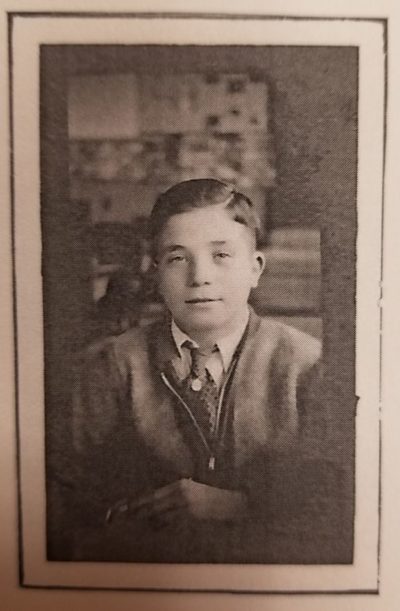 An old, sepia tone photograph of a young boy with his arms crossed. His hair is combed and he wears a tie, shirt and sweater.