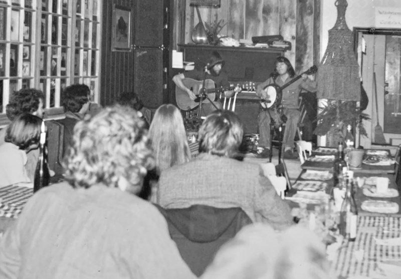 A black and white photograph in which we see the back of audience members as they watch a duo perform music in front of a piano. One plays guitar and the other, a banjo.