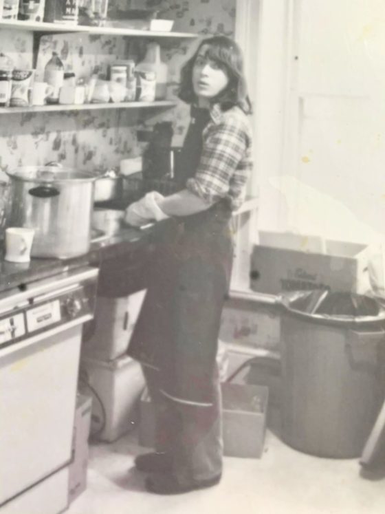 An old black and white photograph. Someone stands in an apron washing dishes in a kitchen. Their hair is shoulder length and they wear rubber gloves and a plaid shirt.