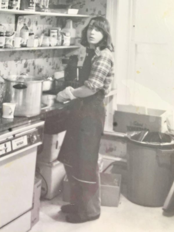 A black and white photograph in which we see a man with long hair and wearing an apron washing dishes in a cafe.