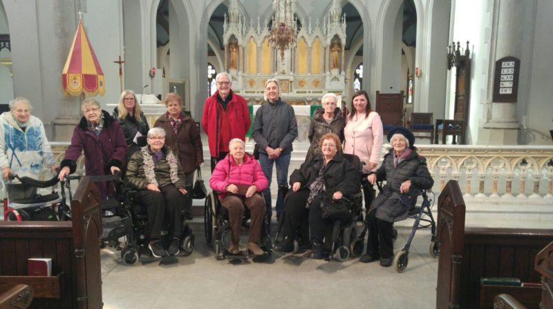 A colour photograph of a group of individuals, some standing, and some in wheelchairs and walkers, wearing jackets in a church.