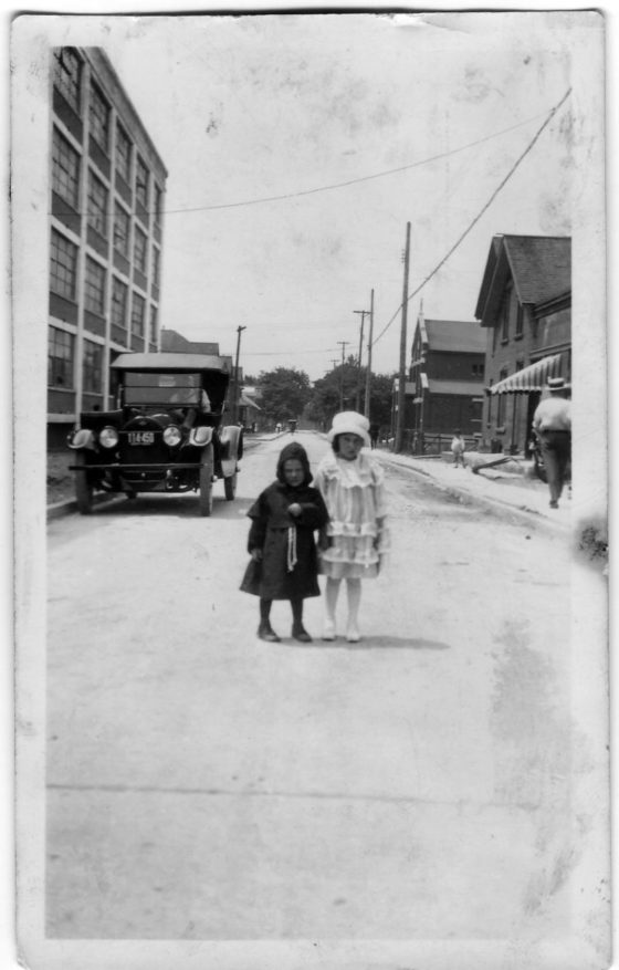 An old black and white photograph. Two young children stand in the middle of a road. They wear dresses and hats.