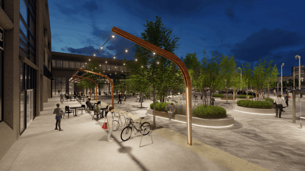 Nighttime scene of a lively urban plaza with string lights hanging from curved wooden poles. People are gathered around tables and walking through the space. Several bicycles are parked near the foreground, and landscaped planters with trees and greenery are lit from below. The surrounding buildings have large glass windows, and the sky is dark.