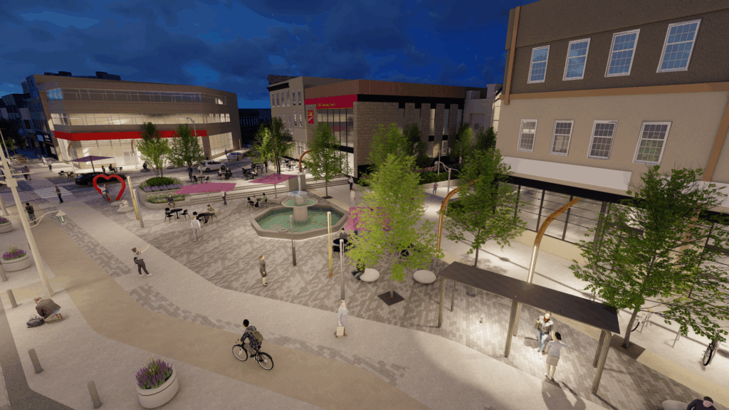 Aerial nighttime view of an urban plaza with a central fountain, purple shade umbrellas, and landscaped areas with trees. People are walking and sitting throughout the space. A large red heart sculpture is visible on the left side, and surrounding streets have minimal traffic. Buildings with modern and traditional architecture frame the plaza, and warm lighting illuminates pathways.