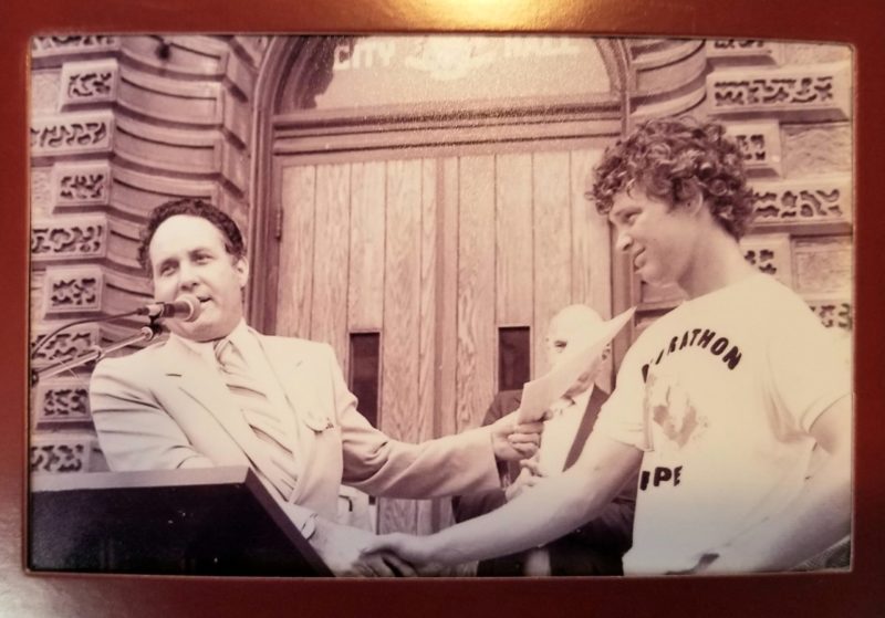 An old, sepia tinted photograph with former Guelph Mayor, Norm Jary, at a podium in front of City Hall, shaking the hand of Terry Fox, who wears a “Marathon of Hope,” t-shirt.