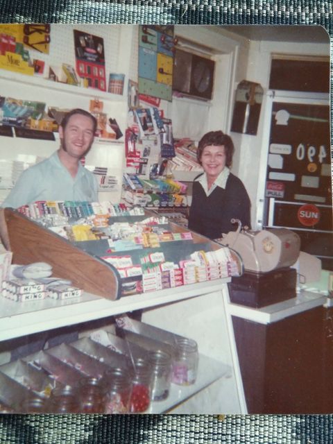 A colour photograph of an old colour photograph. Two people stand behind the counter of a variety shop. They smile for the camera.