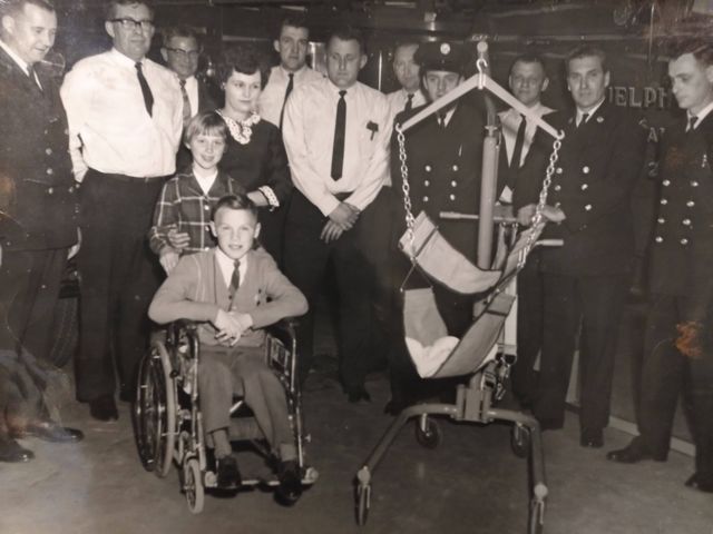 : A black and white photograph of a group of people in formal dress and uniform. A child sits in a wheelchair, next to a swing-seat on wheels.