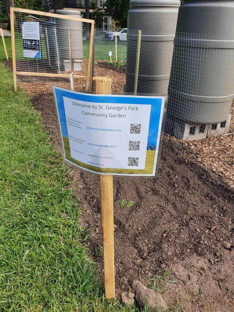 Printed sign reads "Welcome to St. George's Park Community Garden". A fence and rainbarrels are visible behind the sign.