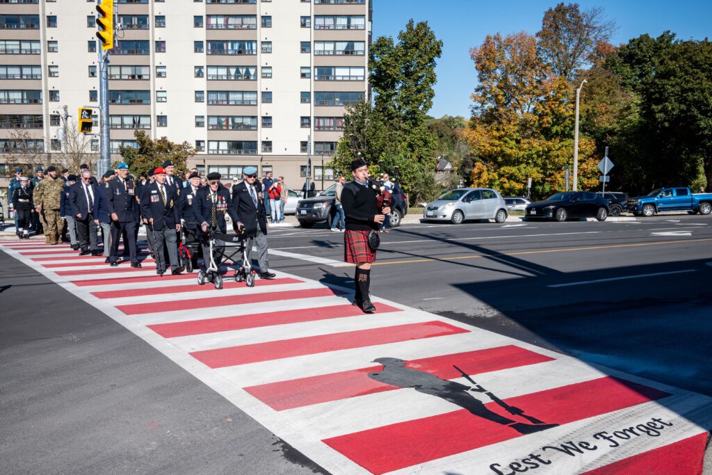 Members of the Royal Canadian Legion cross a red and white striped crosswalk, led by a bagpiper.