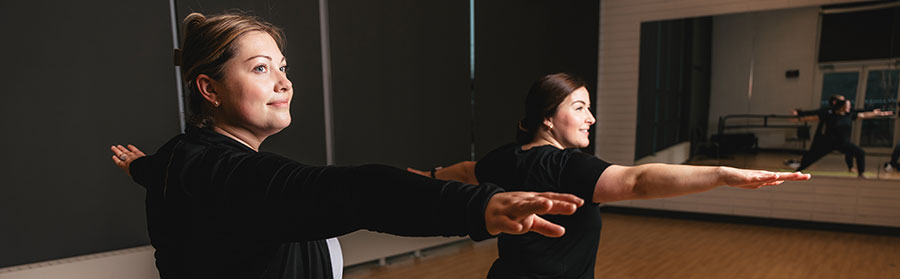 Two women in a fitness studio perform a balancing exercise with their arms extended. They are wearing black workout attire and are focused on their movements. A large mirror reflects their image.