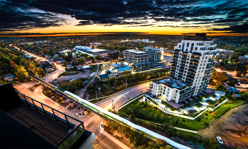 Aerial view of the city of Guelph at the intersection of Woolwich and Macdonell Street. The image shows a dynamic sunset scene over downtown Guelph with city street lights, green space, busy roads and an elevated rail line with the GO train travelling through downtown Guelph with condominium towers in the foreground and City Hall in the background.