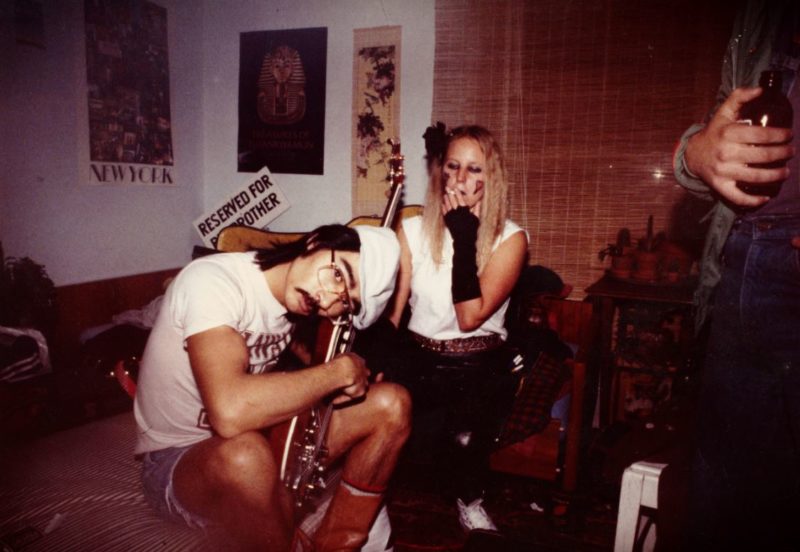 A colour photograph of two individuals on a couch. One has shoulder length hair under a hat and plays guitar, and the other with longer hair, smokes a cigarette.