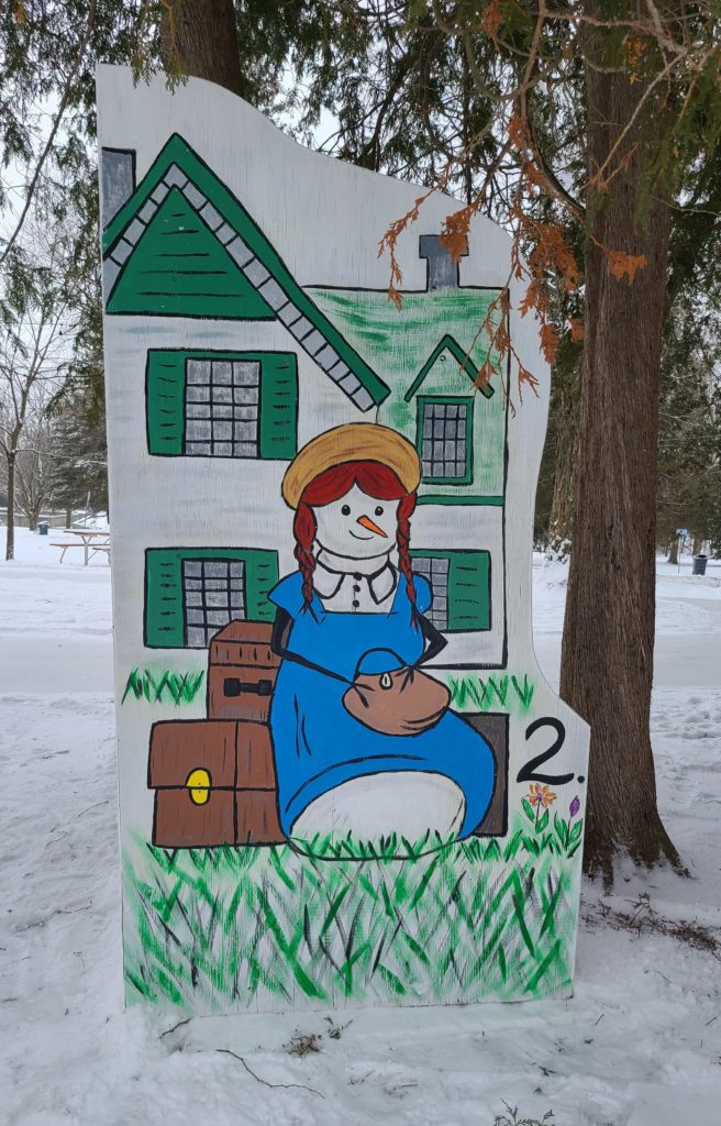 One snowperson with red hair and luggage sits in front of a house with green shutters.