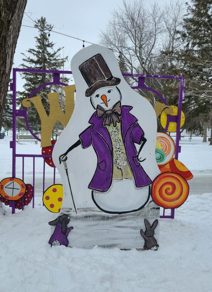 One snowperson in fancy dress poses with a cane in front of gates with “W” on them.