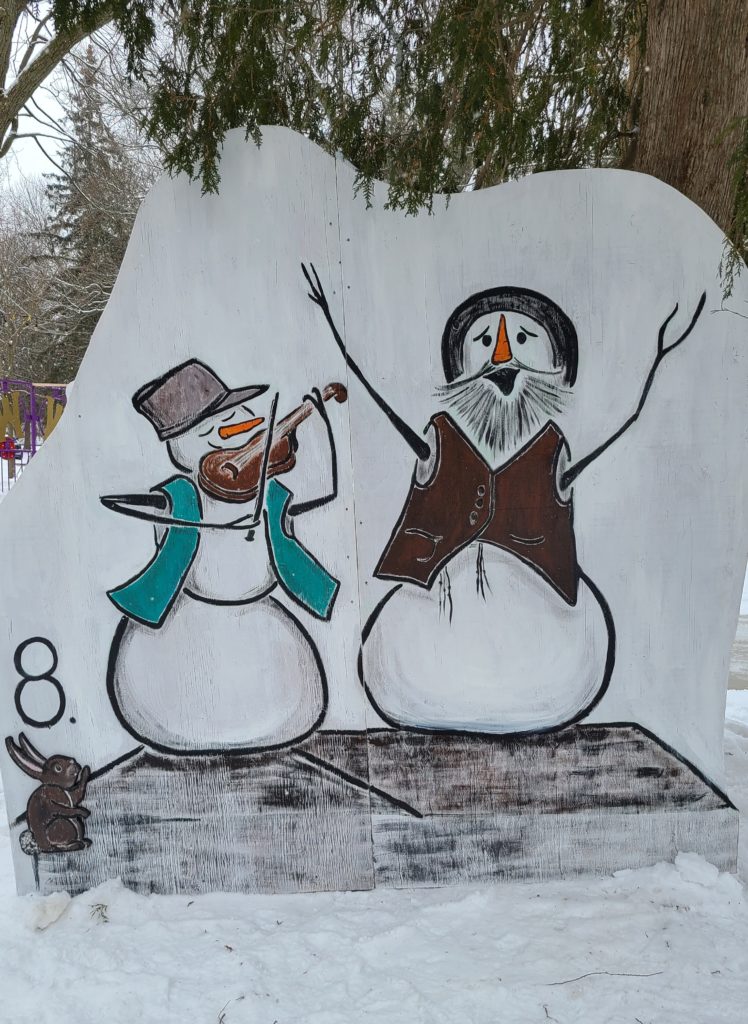 Two snowpeople stand on a roof. Both wear vests and hats. One plays a fiddle while the other sings with arms outstretched.